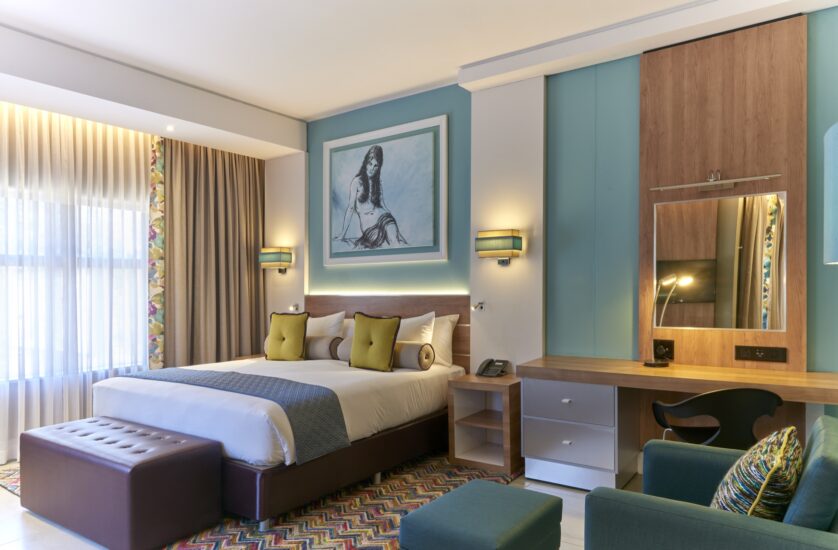 A Junior Suite with teal walls, a framed sketch, work desk and lounge armchair at Metcourt, Emperors Palace