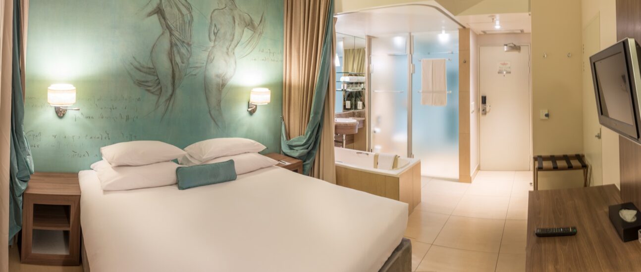A double bed under a figure mural with wall sconces and a glass shower in a Superior Room at Metcourt, Emperors Palace